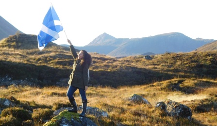 Zoom image: UB Law student standing on a rock holding a flag with mountains in the background in Scotland.