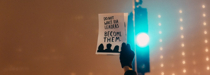 hand holding a sign that reads do not wait for leaders become them. 
