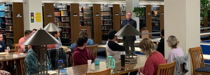 man lecturing to students who are sitting next to a table in a library. 