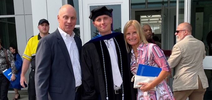 young man (center) wearing graduation regalia, posing with his father and mother, standing outside a building. 