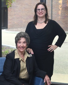two smiling women, one seated, other standing. 