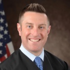 man standing in front of US flag, wearing a judges robe, smiling. 