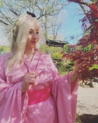 Zoom image: Audrey wearing a pink kimono with a bright pink sash, standing outdoors in a garden. They hold a branch of a red-leafed tree, with sunlight casting leaf-shaped shadows on their clothing. A wooden garden structure and tall trees are visible in the background.