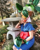 Zoom image: Audrey dressed in a blue outfit with a large red sash, sitting beside a stone lantern in a garden. They hold a prop vegetable with large green leaves, and a basket of similar vegetables is strapped to their head. Fallen autumn leaves and tall trees surround the scene.