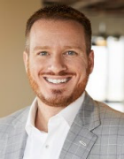 Person with short hair and beard wearing a light gray checkered suit jacket and white dress shirt, posed in an indoor professional setting with a blurred background. 