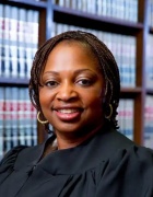 Person wearing a black judicial robe, standing in front of shelves filled with legal books in a law library or office setting. 