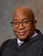 Person wearing glasses and a black judicial-style robe, posed against a neutral background with shades of gray and a hint of red. 