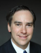 Person wearing a dark suit, white shirt, and striped tie, posed against a plain dark background in a formal setting. 