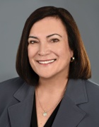 Person with short dark hair wearing a gray blazer over a black top, accessorized with hoop earrings and a circular pendant necklace, posed against a plain gray background. 