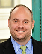 Person wearing dark suit jacket, blue shirt and light green tie, standing in front of an office window. 