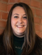 Person with long straight hair wearing a dark green ribbed sweater and geometric necklace, smiling while standing in front of a brick wall. 