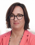 Person with short dark hair wearing glasses and a pink blazer, posed against a blurred white background. 