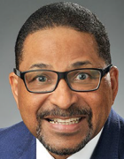 Person wearing glasses and a suit, smiling at the camera against a plain gray background. 