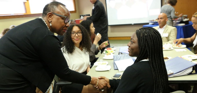 Hon. Stephanie Saunders shakes hands with a participant during a discussion session, while other attendees sit around a conference table. 