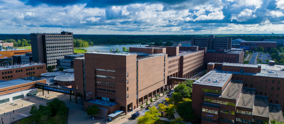 Aerial view of the University at Buffalo North Campus, showing large brick academic buildings, tree lined walkways, parking areas, and a distant lake under a partly cloudy sky.
