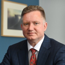 Person wearing a dark plaid suit jacket, white shirt, and red patterned tie, seated indoors with a framed artwork on the wall in the background.&rdquo;. 