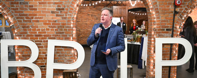 Person speaking into a microphone while standing in front of large white letters arranged in a brick‑walled event space, with attendees seated nearby and additional people gathered in the background.