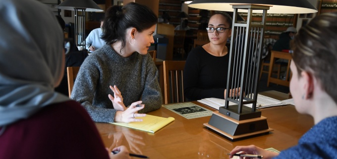 students sitting at a table in a library, talking to a professor. 