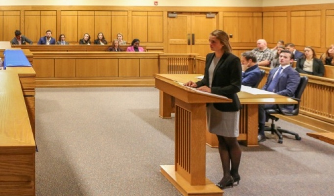 A student stands at a lectern presenting a case in a courtroom while a panel of judges listens. Additional participants sit at tables and in the gallery, observing the mock trial in a wood‑paneled courtroom. 