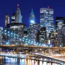 Nighttime city skyline with illuminated skyscrapers and office buildings reflected in a river. A bridge spans the foreground, and blue and white lights from buildings and streets create a bright urban waterfront scene.