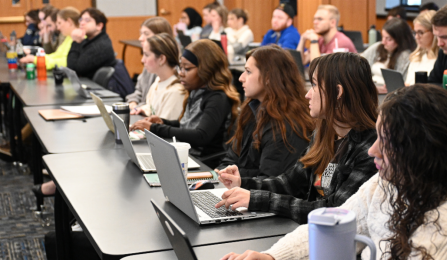 Students sit in rows in a classroom, working on laptops and taking notes during a lecture.