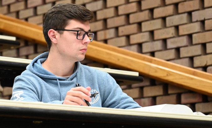 Young man sitting at a desk in a classroom, books in front of him. 
