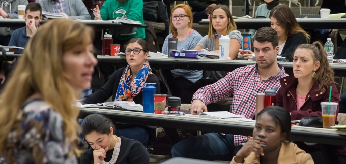 Students seated in a tiered classroom listen to an instructor speaking at the front of the room. 