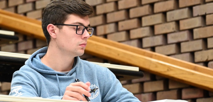 Young man sitting at a desk in a classroom, books in front of him. 