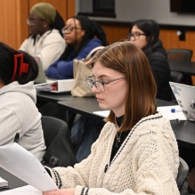 Students seated at tables in a classroom review printed materials and notebooks, with laptops, binders, and water bottles visible on desks. 
