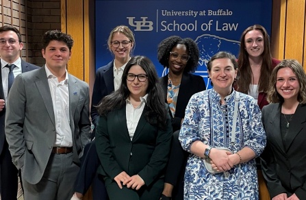 A group of eight individuals dressed in professional attire standing together in front of a blue backdrop that reads “University at Buffalo School of Law” with the UB logo and a buffalo graphic. The setting includes wooden panels and brick walls on either side of the backdrop. The individuals are posed for a group photo, with some standing and others slightly leaning forward.