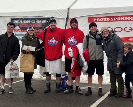 A group of seven people standing together outdoors in front of a large white tent with snow on the ground. The tent has banners that read “Special Olympics New York WELCOME #PlungeNation” and “PROUD SPONSOR.”.