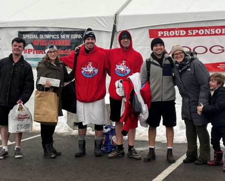 A group of seven people standing together outdoors in front of a large white tent with snow on the ground. The tent has banners that read “Special Olympics New York WELCOME #PlungeNation” and “PROUD SPONSOR.”. 