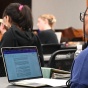 A classroom scene with several students seated at tables using laptops. The focus is on a student in the foreground wearing glasses and working on a laptop that displays a document. Other students sit at separate tables facing the front of the room, where a large screen is mounted on the wall. 