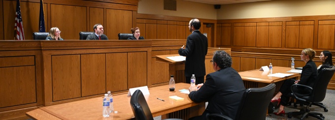 Courtroom scene with a panel of three judges seated behind a wooden bench while an attorney stands and speaks at a podium. Additional attorneys sit at tables facing the judges, with water bottles and papers arranged on the desks.
