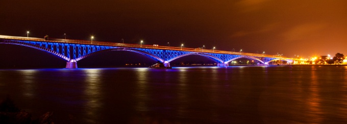 Nighttime view of a long bridge illuminated with vibrant blue lights, reflecting off the dark water below, with an orange-glowing sky and distant city lights in the background. 