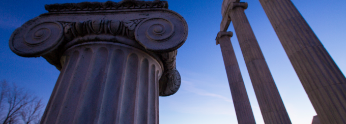 Upward view of large ancient-style columns with ornate capitals, set against a deep blue sky. One column is close in the foreground, and several taller columns with a partial entablature appear in the background. 