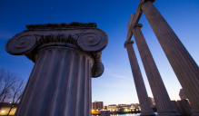 Upward view of large ancient-style columns with ornate capitals, set against a deep blue sky. One column is close in the foreground, and several taller columns with a partial entablature appear in the background. 