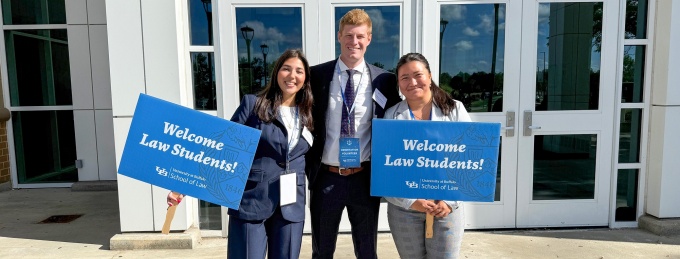 Three University at Buffalo School of Law volunteers stand outside a campus building entrance holding blue signs that read, “Welcome Law Students!”. 