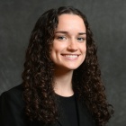 Professional portrait of Calprin, long brown curly hair, wearing a black jacket and and top. 