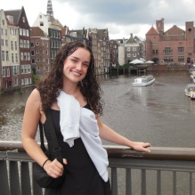 Person standing on a bridge overlooking a canal lined with narrow buildings. Boats are visible on the water below, and the surrounding architecture features tall, closely spaced facades under a cloudy sky, indicating an urban waterfront setting. 
