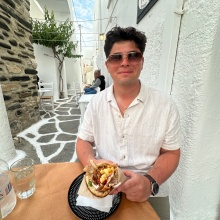 A person sits at an outdoor caf&eacute; table in a narrow, white‑washed alley with stone‑patterned pavement. They are holding a plate with a pita sandwich filled with vegetables and meat. Glasses of water and tableware are on the table, and other diners are seated further down the walkway. 