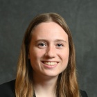 Professional headshot of a person with long hair wearing a black blazer and light-colored top, seated against a gray studio backdrop and looking toward the camera. 