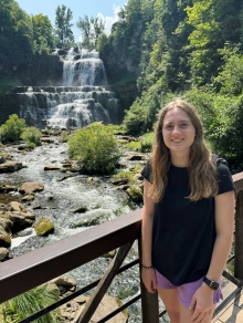 Person standing on a metal pedestrian bridge in front of a multi tiered waterfall surrounded by trees and rocks, with a stream flowing below on a sunny day. 