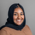 A person wearing a black headscarf and a brown buttoned sweater poses for a studio portrait against a plain gray background. 