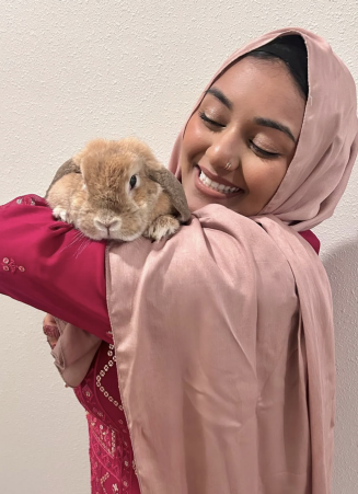 A person wearing a light pink headscarf and a bright pink patterned outfit holds a light brown rabbit resting on their arm. The background is a plain, light‑colored wall. 