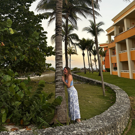 woman smiling, standing outside, hugging a palm tree. 