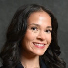 A person with long dark hair wears a dark navy blazer over a white top and a small pendant necklace while posing for a studio portrait against a dark textured background. 
