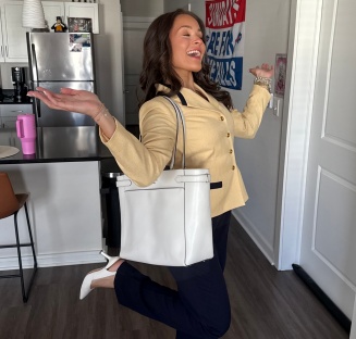 Person standing in an apartment entryway wearing a light-colored blazer, dark pants, and white shoes, carrying a large white tote bag over one shoulder. One leg is raised while both arms are extended outward. A kitchen with stainless steel appliances is visible in the background.