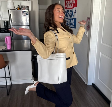 Person standing in an apartment entryway wearing a light-colored blazer, dark pants, and white shoes, carrying a large white tote bag over one shoulder. One leg is raised while both arms are extended outward. A kitchen with stainless steel appliances is visible in the background. 