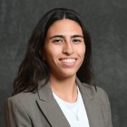 A person with long dark hair wears a light gray blazer over a pale top and a small pendant necklace while posing for a studio portrait against a dark textured background. 